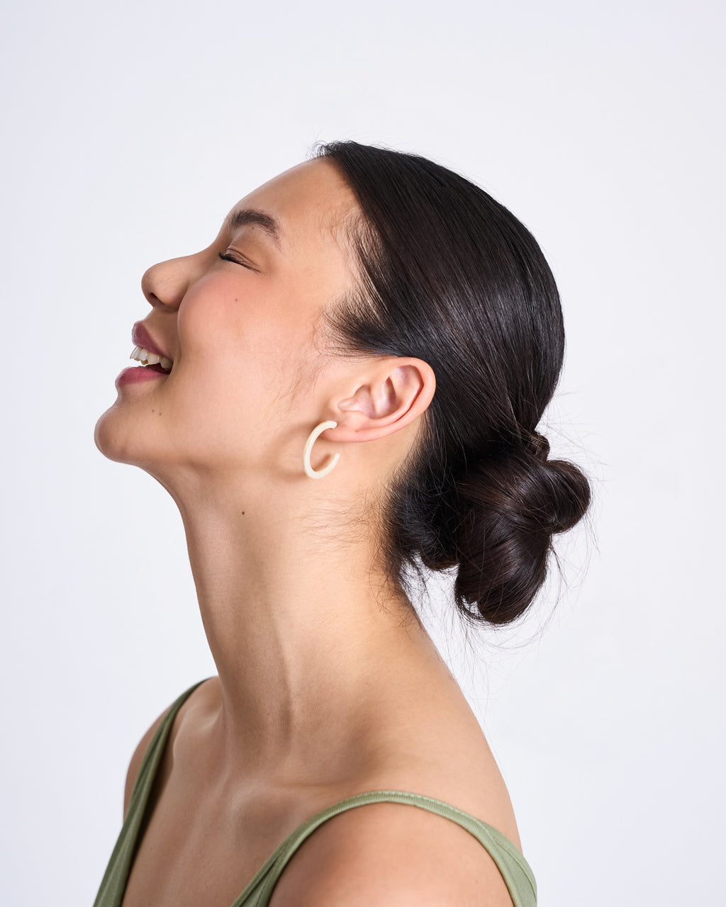 A woman with dark hair in a low bun, wearing a green top and jan-n-june MINI HOOPS IVORY earrings, smiles with her eyes closed against a plain light background.