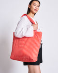 A woman stands against a plain light background, carrying the jan-n-june PADDED SHOPPER SMOL in cherry red with white stripes over her shoulder, styled with a white blouse and black shorts.