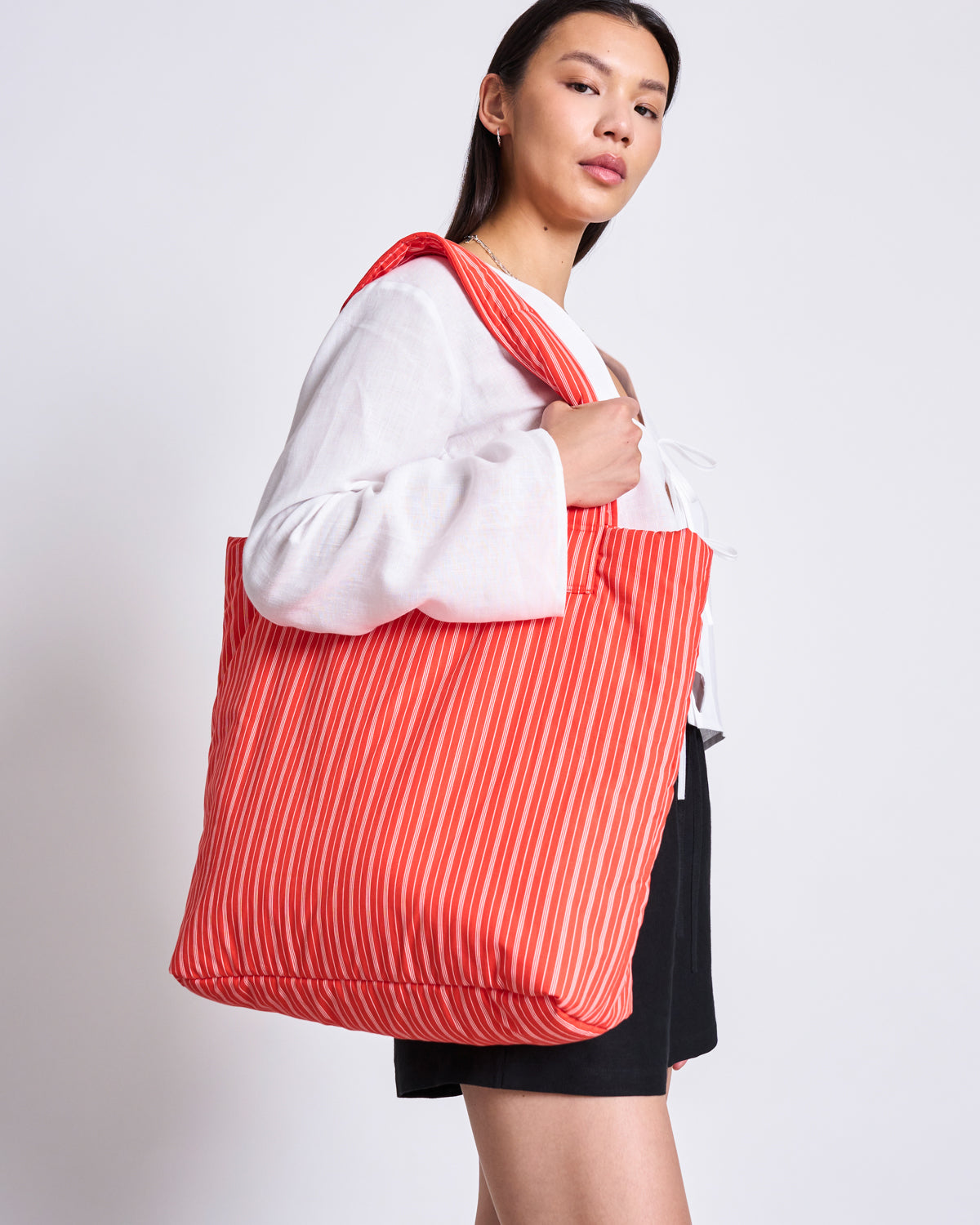 A woman stands against a plain light background, carrying the jan-n-june PADDED SHOPPER SMOL in cherry red with white stripes over her shoulder, styled with a white blouse and black shorts.