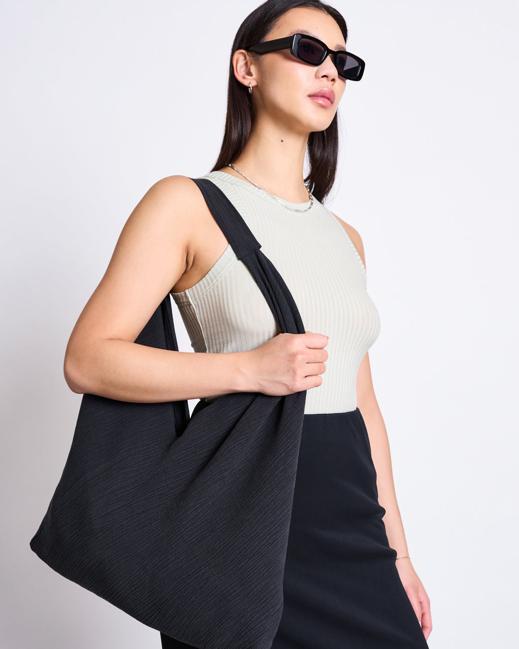 A woman in black sunglasses, a sleeveless beige ribbed top, and a black skirt stands against a white background, holding the spacious jan-n-june CRINKLE SHOPPER VISBY BLACK made from Tencel™ Lyocell.