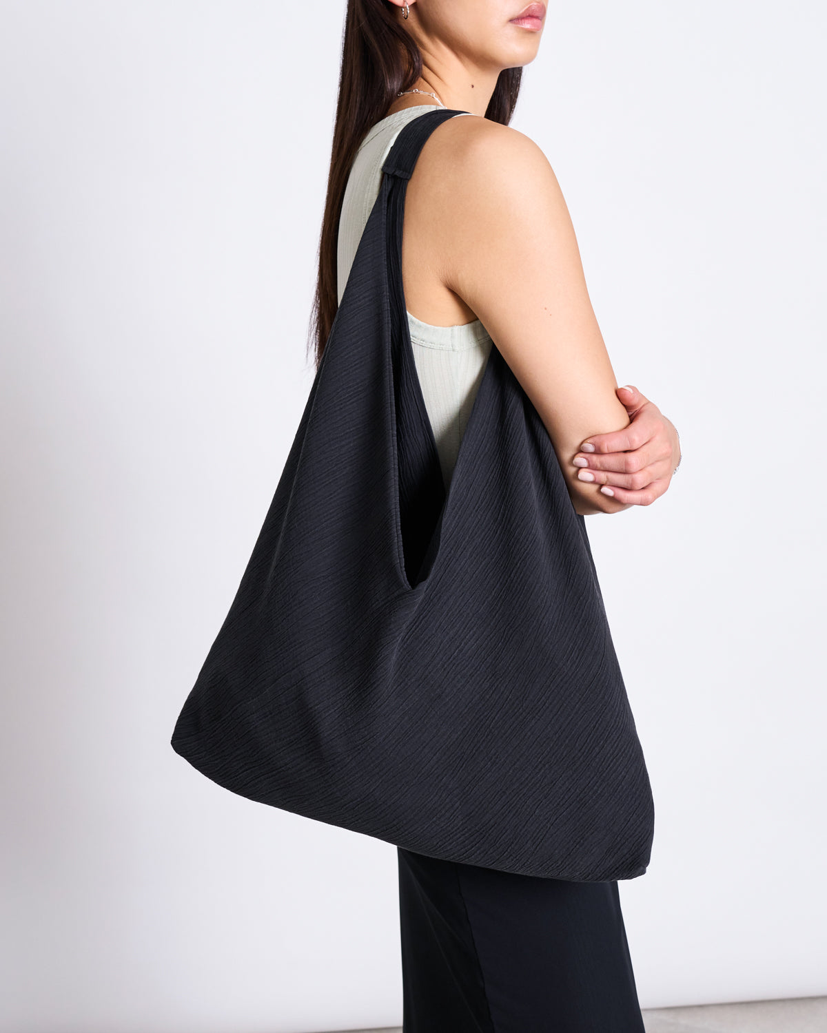 A woman with long hair stands sideways, holding the jan-n-june CRINKLE SHOPPER VISBY BLACK—a large, slouchy shoulder bag—over her shoulder. She wears a sleeveless light top and black pants against a plain white background.