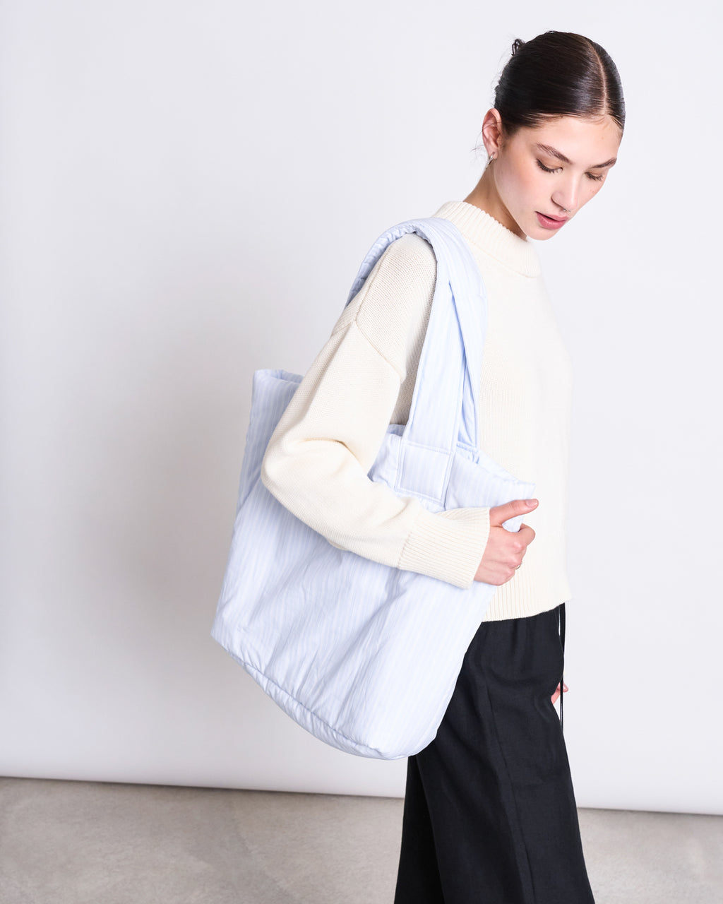 A woman with dark hair tied back wears a cream sweater and black pants, looking down while carrying the jan-n-june PADDED SHOPPER SMOL BLUE WHITE STRIPED over her shoulder against a plain background.
