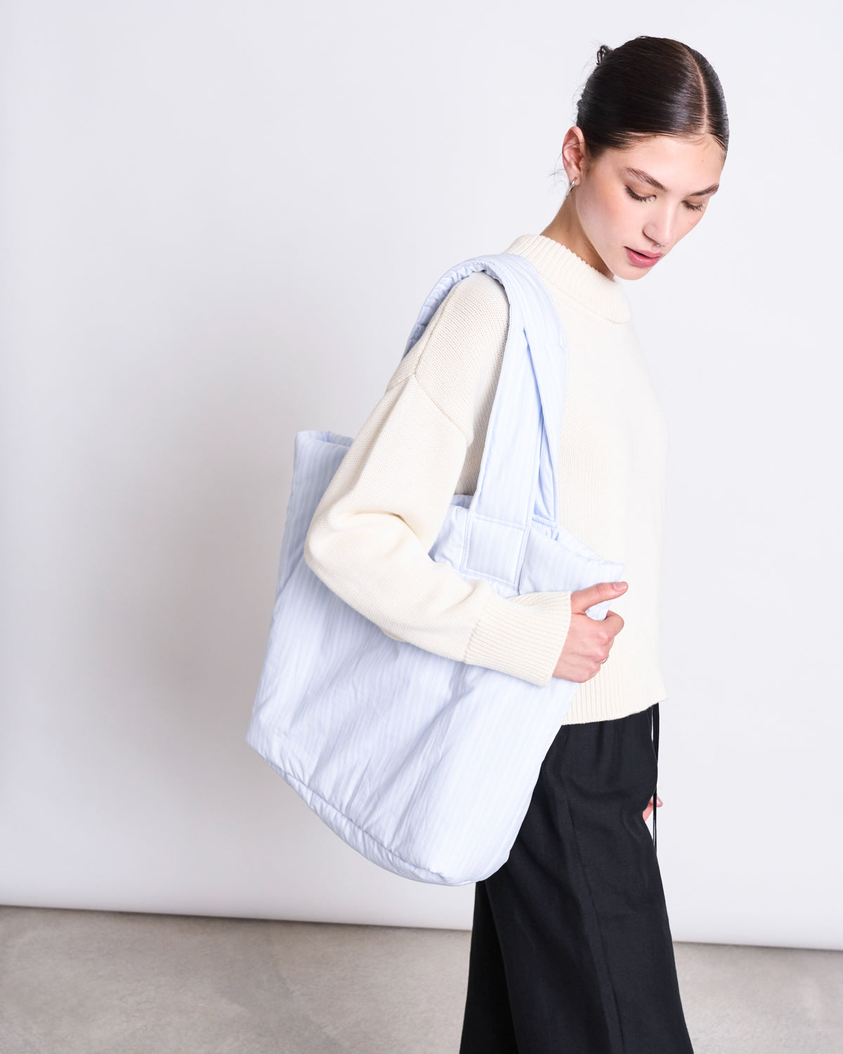 A woman with dark hair tied back wears a cream sweater and black pants, looking down while carrying the jan-n-june PADDED SHOPPER SMOL BLUE WHITE STRIPED over her shoulder against a plain background.