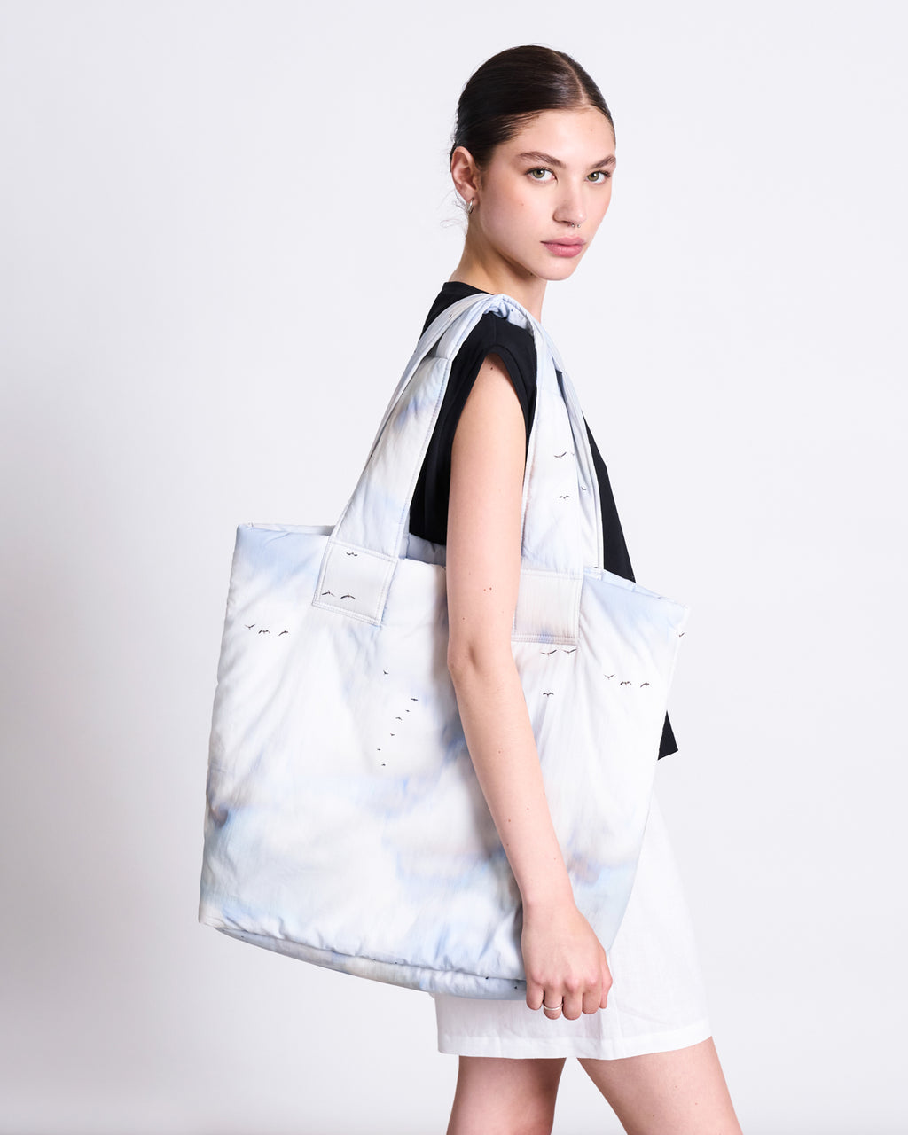 A woman with dark hair in a low bun wears a black top and white shorts, carrying the jan-n-june PADDED SHOPPER SMOL CLOUD PRINT, a large organic cotton bag with a subtle cloud pattern, over her shoulder against a plain light background.