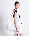 A woman with dark hair in a low bun wears a black top and white shorts, carrying the jan-n-june PADDED SHOPPER SMOL CLOUD PRINT, a large organic cotton bag with a subtle cloud pattern, over her shoulder against a plain light background.