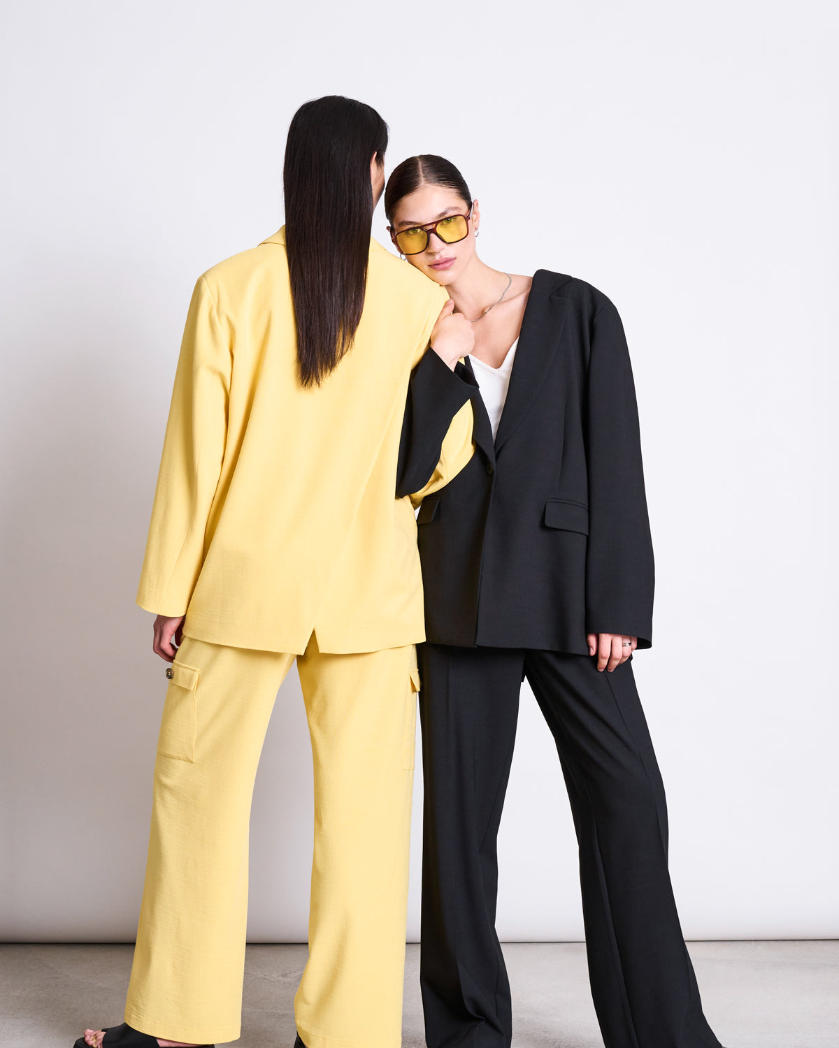 Two women pose together; one wears a yellow pantsuit and faces away, while the other, in jan-n-junes SLITTED BACK BLAZER JELS BUTTERCUP with sunglasses, faces forward and links arms with her.