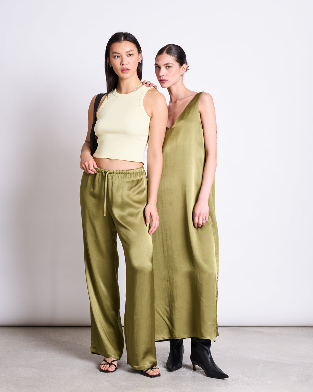 Two women stand against a white wall. One wears a pale yellow crop top with jan-n-june LOOSE SILKY PANTS FRIS WOODY OLIVE and sandals, while the other wears an olive green vegan silk dress with black boots.