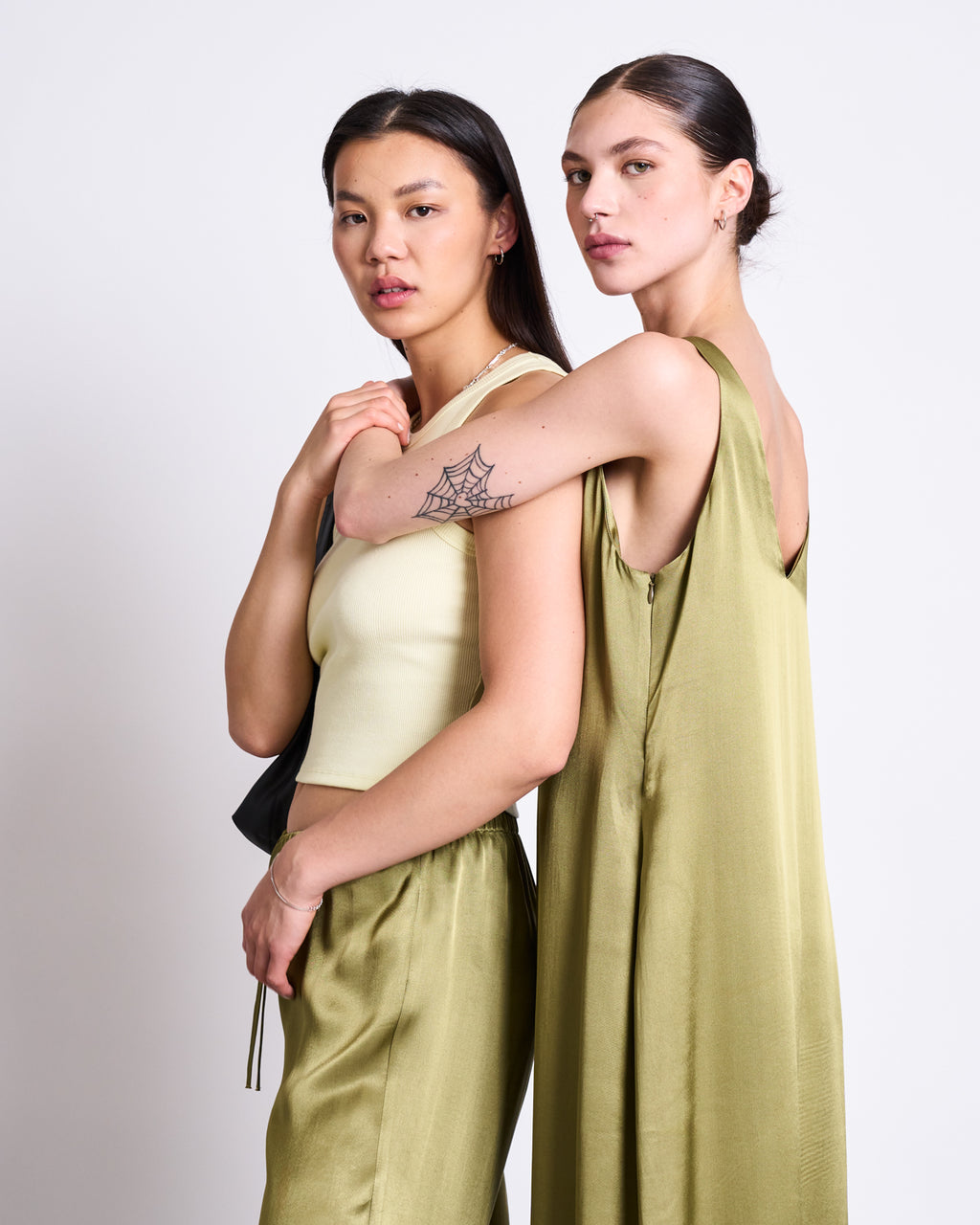 Two women stand closely together against a plain backdrop. One wears a sleeveless cream top with light green pants; the other wears the jan-n-june MAXI DRESS BENVIE WOODY OLIVE, revealing a spider web tattoo on her arm. Both face the camera.