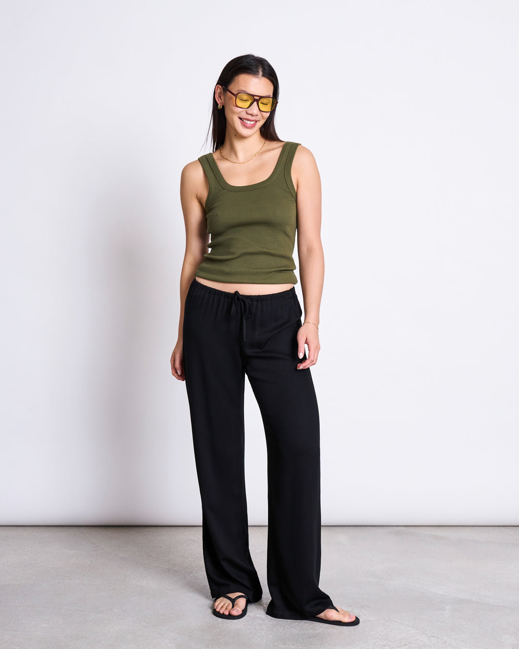A woman stands against a plain white wall, smiling with her hands by her sides. She wears jan-n-junes MID RISE LOOSE PANTS STRATH BLACK, styled with a sleeveless olive green tank top, black sandals, and yellow-tinted sunglasses.