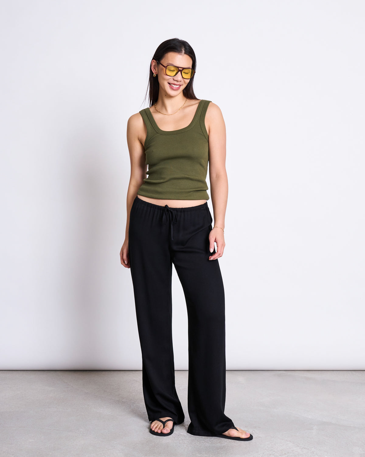 A woman stands against a plain white wall, smiling with her hands by her sides. She wears jan-n-junes MID RISE LOOSE PANTS STRATH BLACK, styled with a sleeveless olive green tank top, black sandals, and yellow-tinted sunglasses.