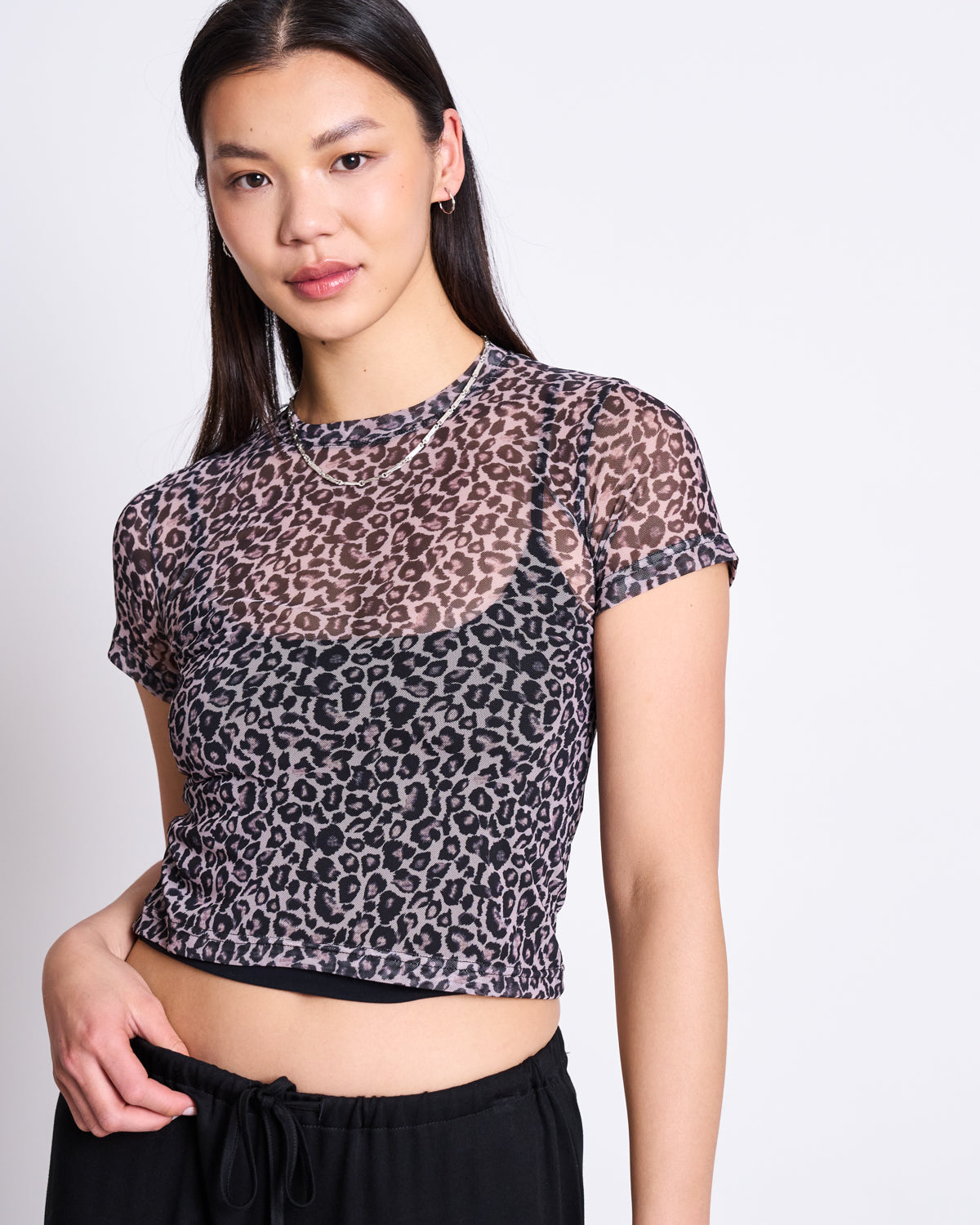 A woman with long dark hair wears the jan-n-june MESH T-SHIRT EVIE LEO PRINT over a black bra and drawstring pants, standing against a light background—an ideal choice for bold festival style.