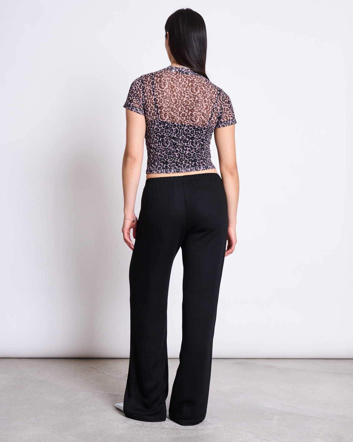 A woman with long dark hair faces away, wearing the jan-n-june MESH T-SHIRT EVIE LEO PRINT and loose black pants on a light gray floor against a white background—an ideal pick for festival style.