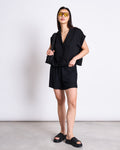 A woman with straight dark hair poses confidently against a plain background, wearing yellow sunglasses, a black short-sleeve button-up shirt, jan-n-june DRAWSTRING SHORTS LOV BLACK, and black slide sandals.