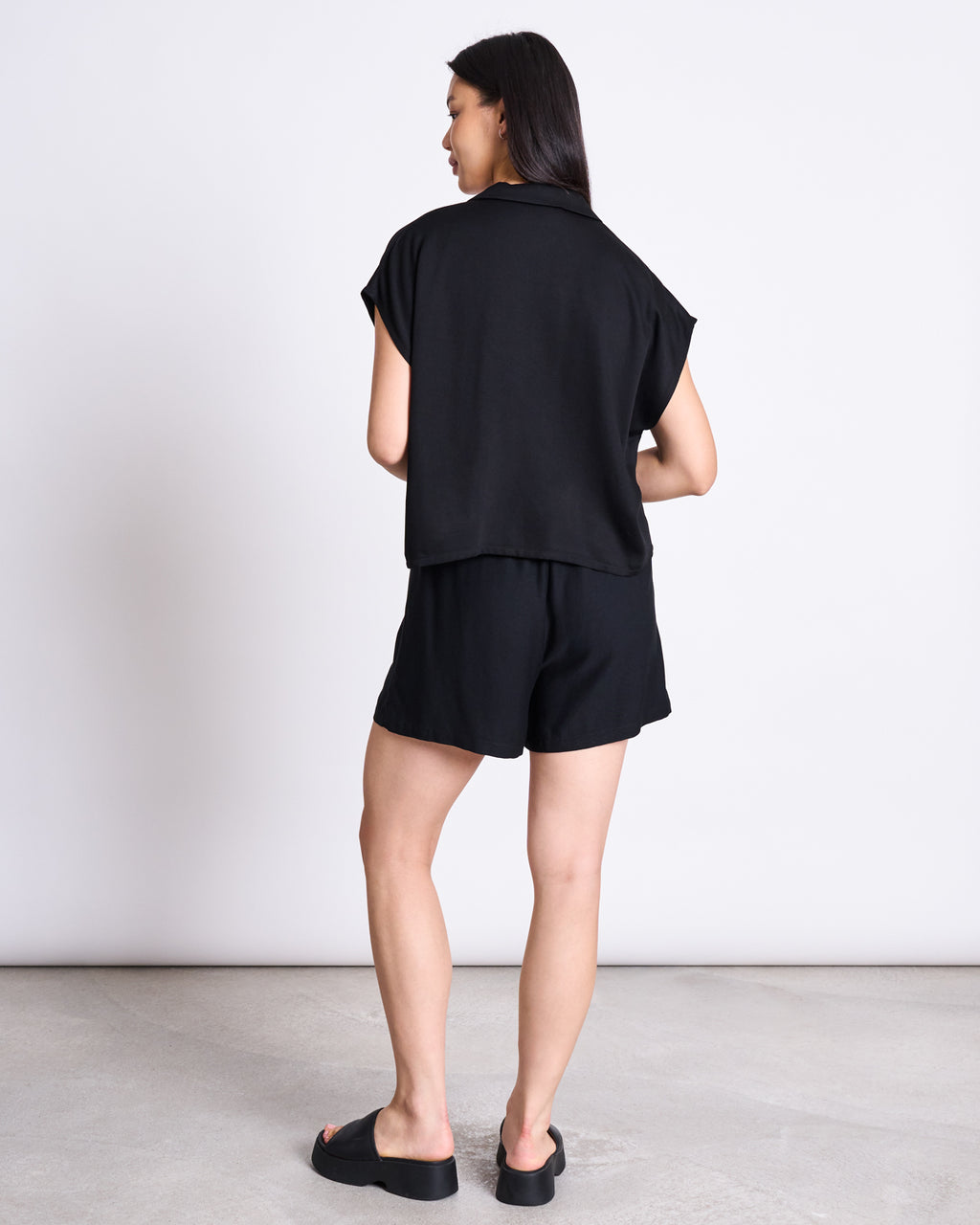 A woman with long dark hair faces away, wearing jan-n-junes DRAWSTRING SHORTS LOV BLACK set—a loose black short-sleeve top and EcoVero™ drawstring shorts—with black slide sandals, standing on a light floor against a plain background.