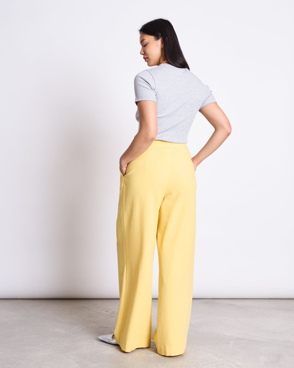 A woman faces away from the camera, wearing jan-n-junes PLEATED WIDE PANTS NIVA BUTTERCUP in yellow and a light gray short-sleeve top. She stands with hands in her pockets on a light floor against a white background.
