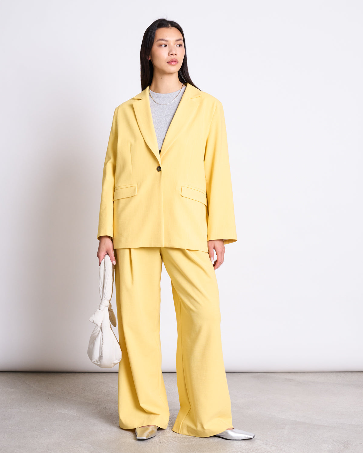 A woman poses in the jan-n-june SLITTED BACK BLAZER JELS BUTTERCUP, featuring an oversized yellow suit with wide-leg pants, a slitted back blazer, a light gray top, silver shoes, and a white knotted handbag.