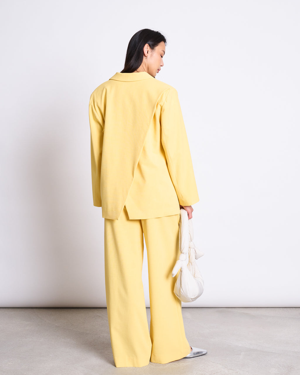 A woman faces away, wearing the jan-n-june SLITTED BACK BLAZER JELS BUTTERCUP—a loose yellow suit with wide-leg trousers and an oversized blazer—holding a white bag against a plain white backdrop.