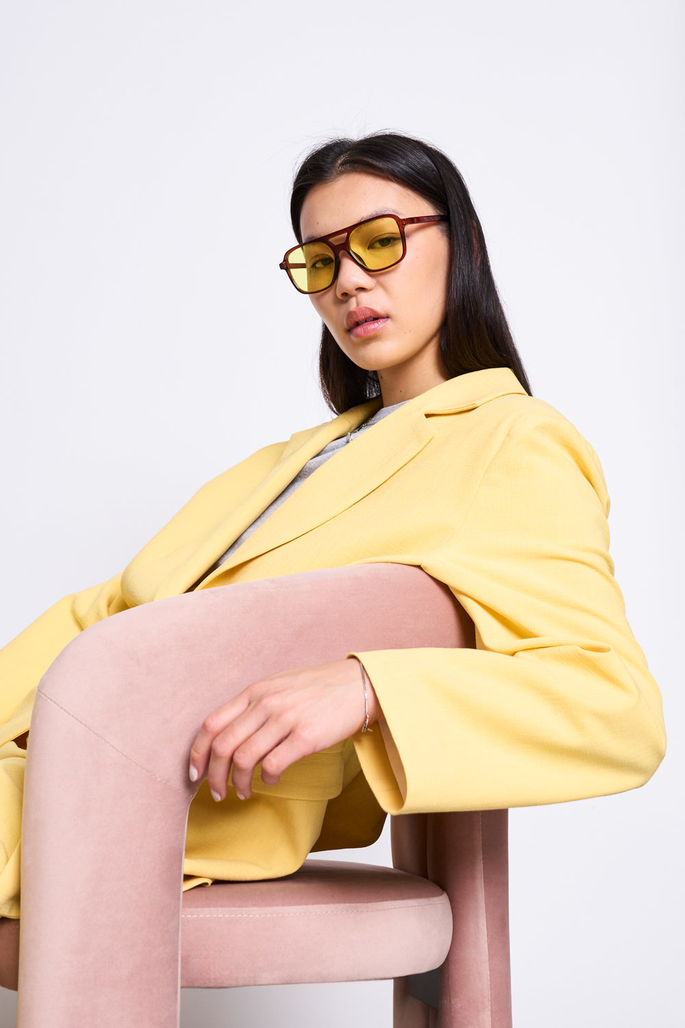 A woman wears the jan-n-june SLITTED BACK BLAZER JELS BUTTERCUP and yellow-tinted sunglasses, sitting confidently on a pale pink chair and looking at the camera against a plain white background.
