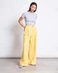 A woman stands against a plain white wall, wearing jan-n-junes PLEATED WIDE PANTS NIVA BUTTERCUP in yellow with a light gray fitted tee and gray pointed-toe shoes, looking down with her hands in her pockets.