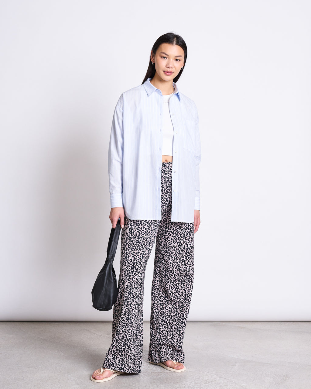 A woman stands against a white background wearing the jan-n-june OVERSIZED SHIRT MAMRO BLUE WHITE STRIPED GOTS over a white crop top, with patterned wide-leg pants, flip-flops, and holding a black bag.