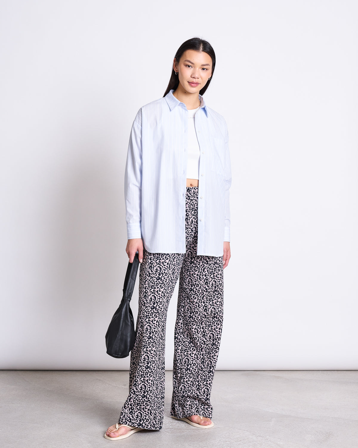 A woman stands against a white background wearing the jan-n-june OVERSIZED SHIRT MAMRO BLUE WHITE STRIPED GOTS over a white crop top, with patterned wide-leg pants, flip-flops, and holding a black bag.