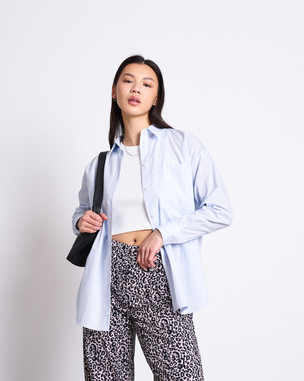 A woman poses confidently against a white background, wearing the jan-n-june OVERSIZED SHIRT MAMRO BLUE WHITE STRIPED GOTS over a white crop top with leopard print pants and carrying a black shoulder bag.
