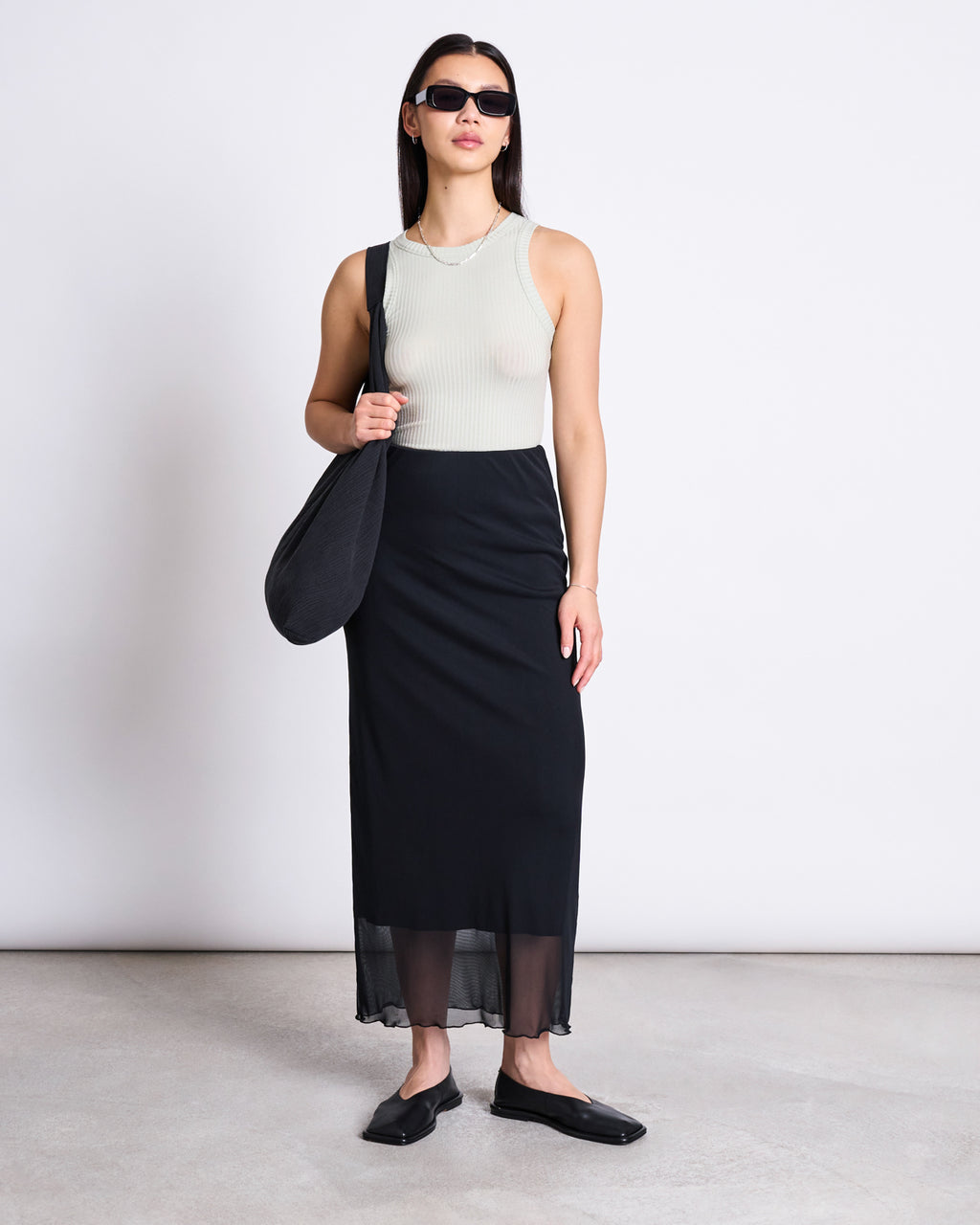 A woman wearing sunglasses, the jan-n-june LYOCELL RIB TANK TOP PORTO VINTAGE LACE, a long black skirt, black flat shoes, and a black shoulder bag stands against a white background.