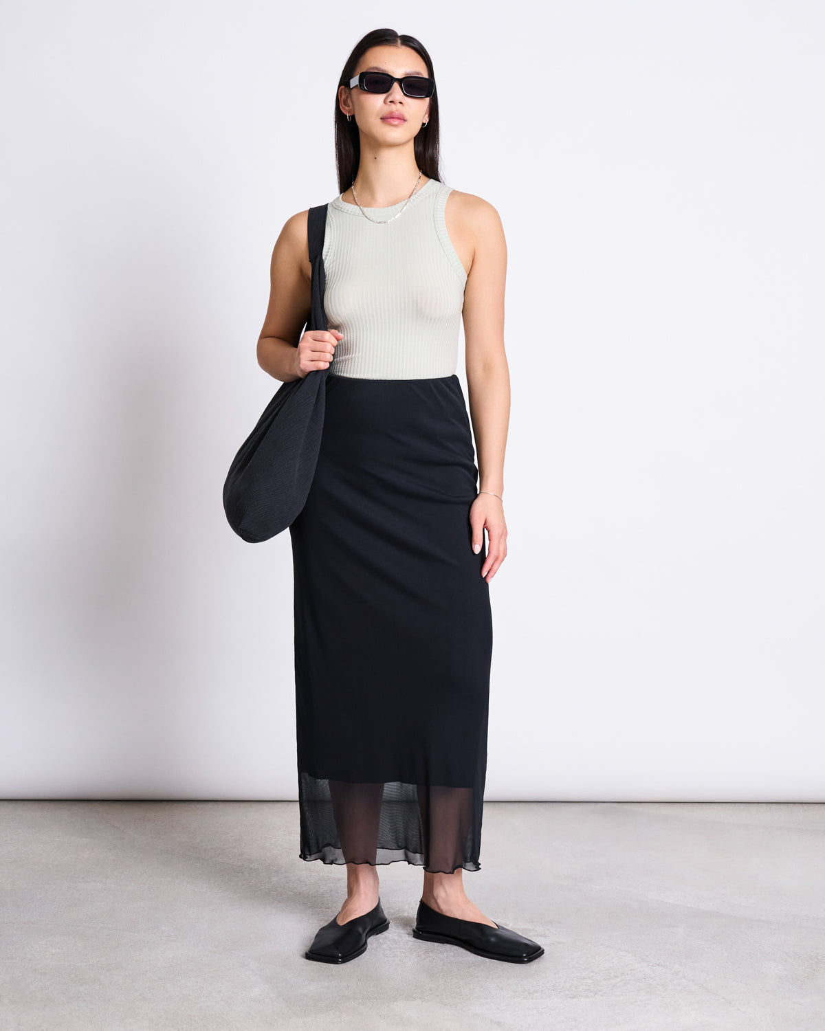 A woman wearing sunglasses, the jan-n-june LYOCELL RIB TANK TOP PORTO VINTAGE LACE, a long black skirt, black flat shoes, and a black shoulder bag stands against a white background.
