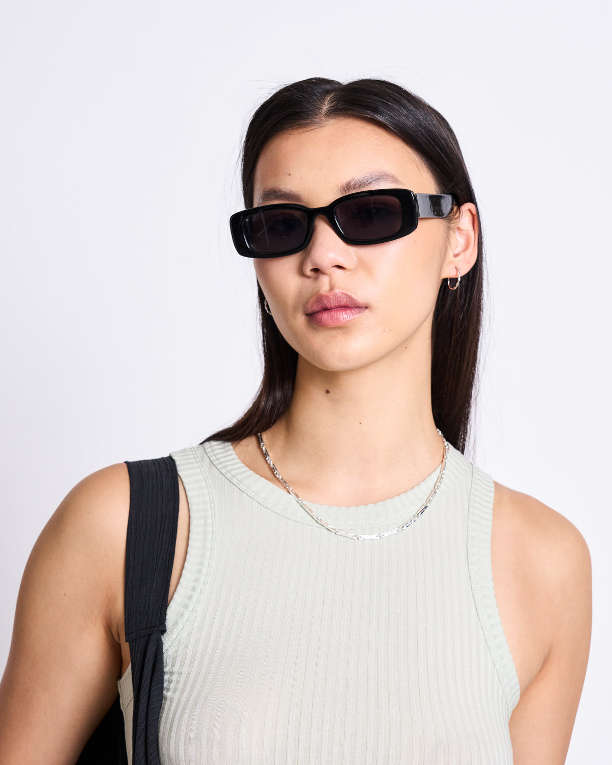 A woman with long dark hair wears black rectangular sunglasses, a silver chain necklace, and carries a black bag, styled in the LYOCELL RIB TANK TOP PORTO VINTAGE LACE by jan-n-june against a plain white background.