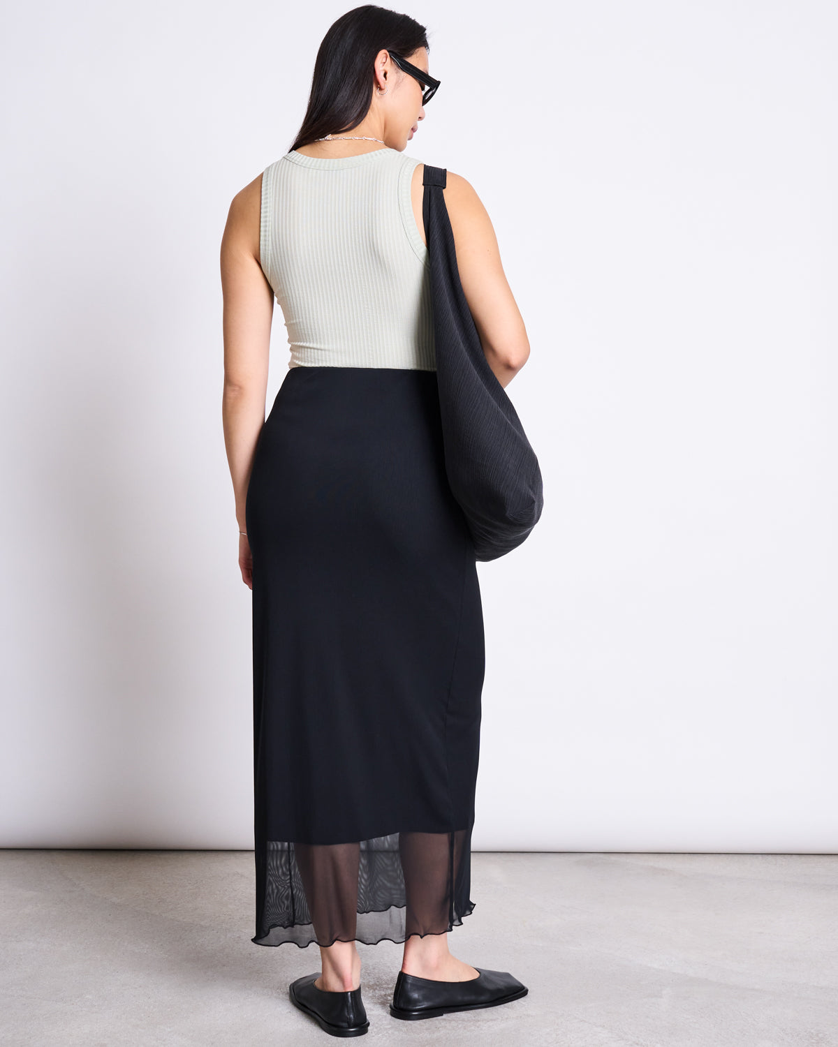 A woman with long dark hair wears the jan-n-june MESH MAXI SKIRT SMUK BLACK, a sleeveless ribbed top, black flats, dark sunglasses, and carries a large black shoulder bag. She stands facing away on a plain background.