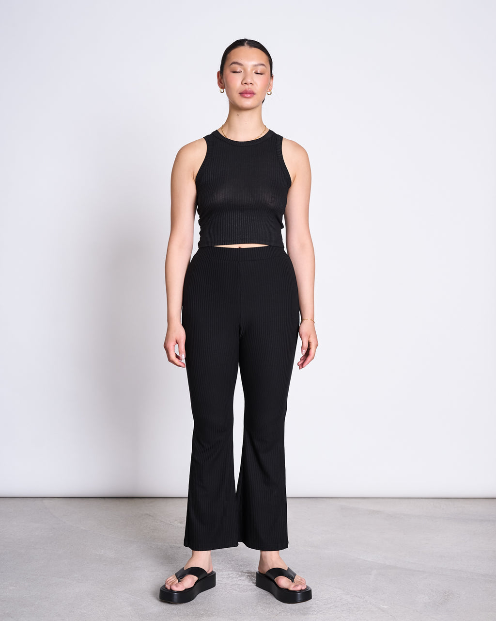 A woman wears the jan-n-june LYOCELL RIB TANK TOP PORTO BLACK and matching high-waisted Porto black pants with slight flares, styled with black platform sandals against a plain white wall.