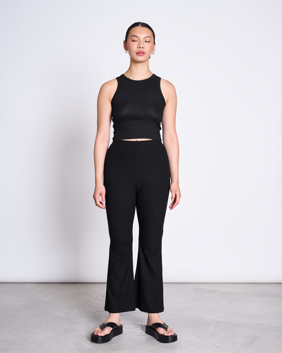 A woman wears the jan-n-june LYOCELL RIB TANK TOP PORTO BLACK and matching high-waisted Porto black pants with slight flares, styled with black platform sandals against a plain white wall.
