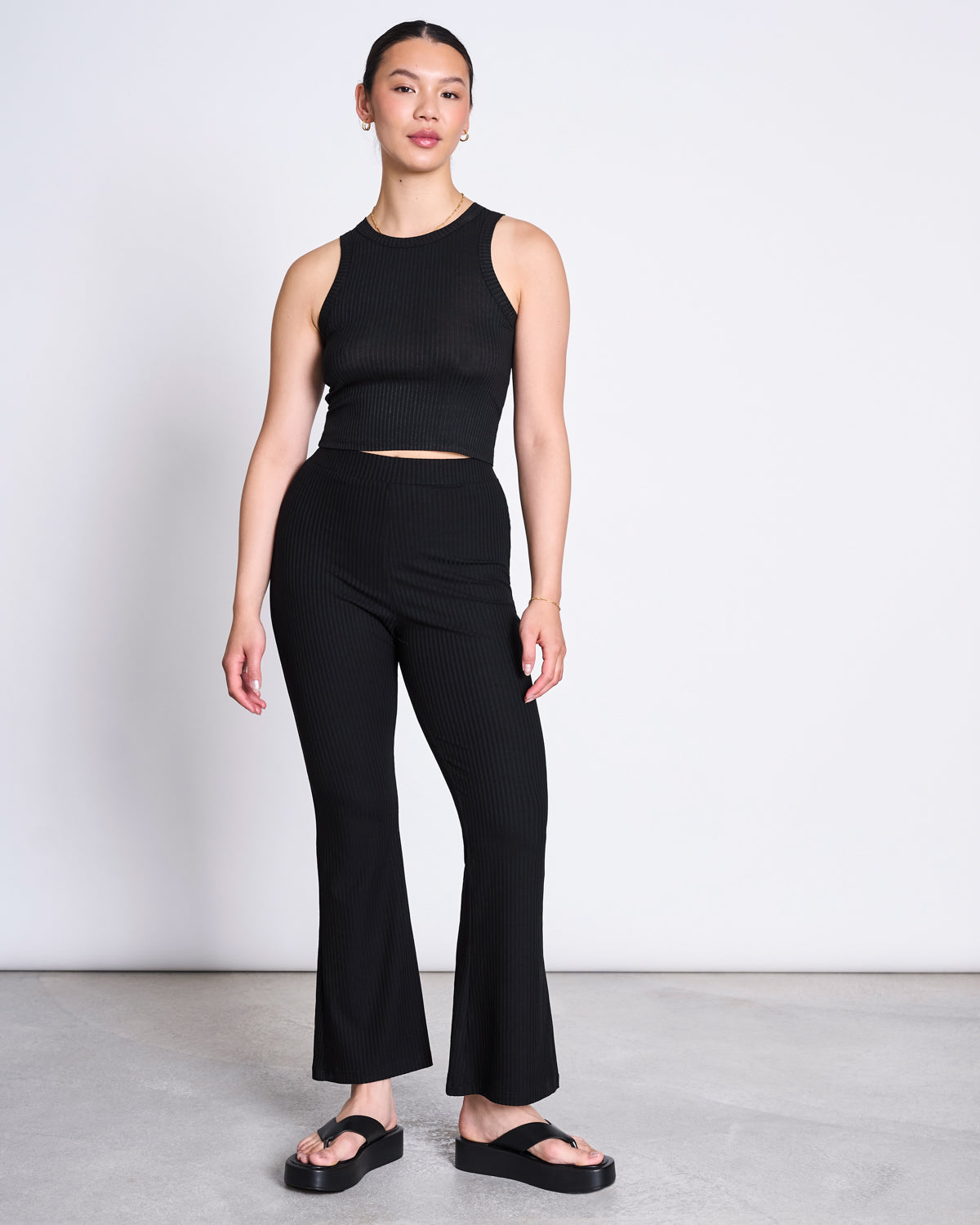 A woman stands against a plain background wearing the jan-n-june LYOCELL RIB TANK TOP PORTO BLACK, paired with high-waisted black pants and black slide sandals; she faces forward with one arm relaxed and the other slightly bent.