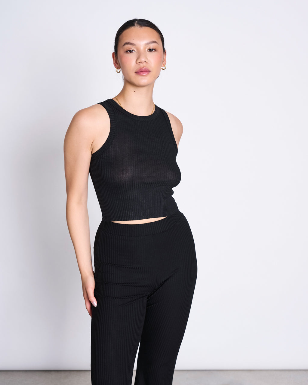 A woman with dark hair tied back wears the jan-n-june LYOCELL RIB TANK TOP PORTO BLACK, a sleeveless, fitted crop top made from Tencel™ Lyocell rib fabric, paired with matching high-waisted pants against a plain white background.