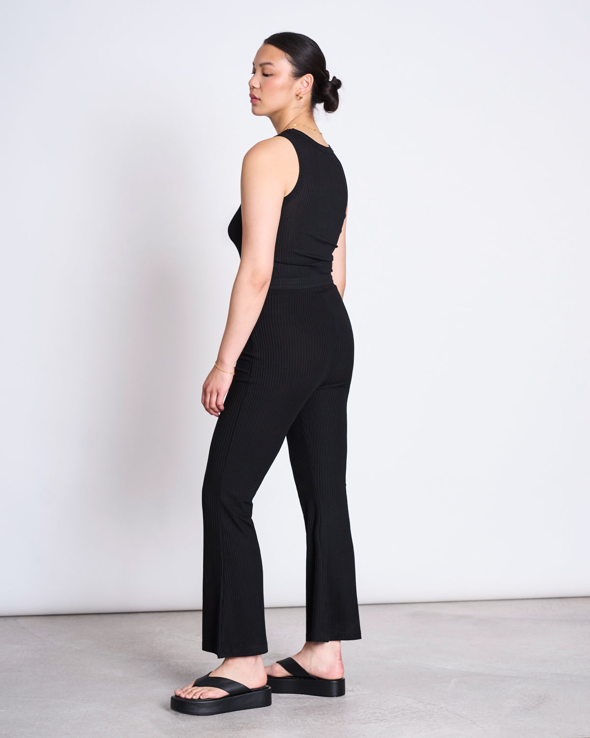 A woman with dark hair in a bun stands sideways, looking over her shoulder. She wears the jan-n-june LYOCELL RIB TANK TOP PORTO BLACK and matching pants, both in soft Tencel™ Lyocell rib fabric, with black platform sandals on a light gray floor.