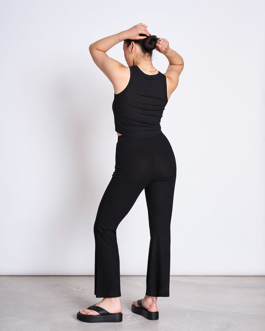 A person stands with arms raised, wearing the jan-n-june LYOCELL RIB TANK TOP PORTO BLACK, black flared pants, and black platform sandals against a plain light background.