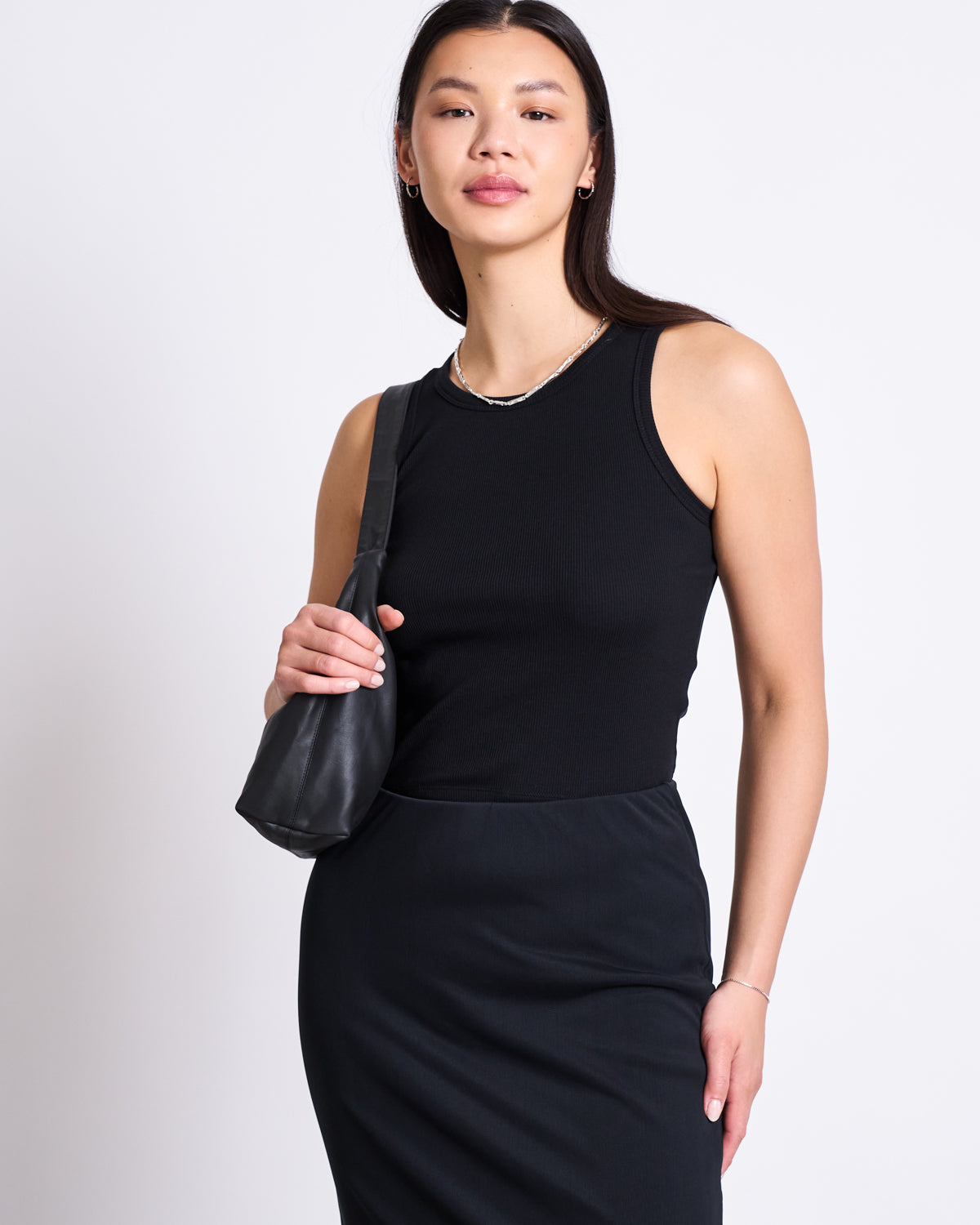 A woman with long dark hair wears the jan-n-june MESH MAXI SKIRT SMUK BLACK and a sleeveless black top, standing against a white background while holding a small black shoulder bag and smiling softly.