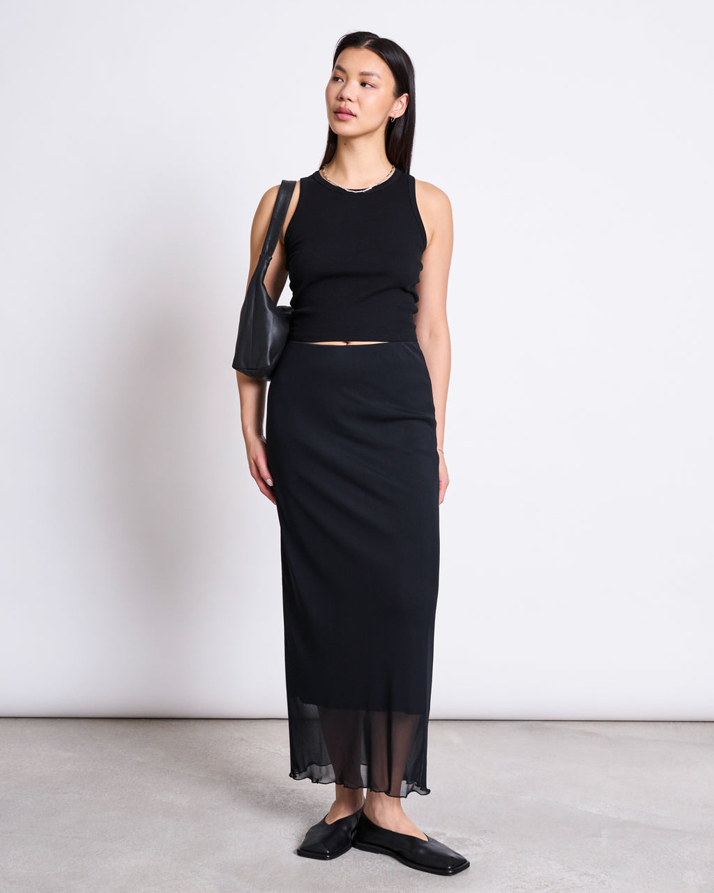 A woman stands against a white wall wearing the jan-n-june TRANSPARENT BACK TANK TOP HOLMEN BLACK, paired with a long black skirt with a sheer hem, black flats, and a black shoulder bag. She looks to the side with a neutral expression.