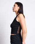 A woman stands in profile against a plain background, wearing the TRANSPARENT BACK TANK TOP HOLMEN BLACK by jan-n-june with mesh sides and black pants. Her straight dark hair is down, and her arms are relaxed at her sides.