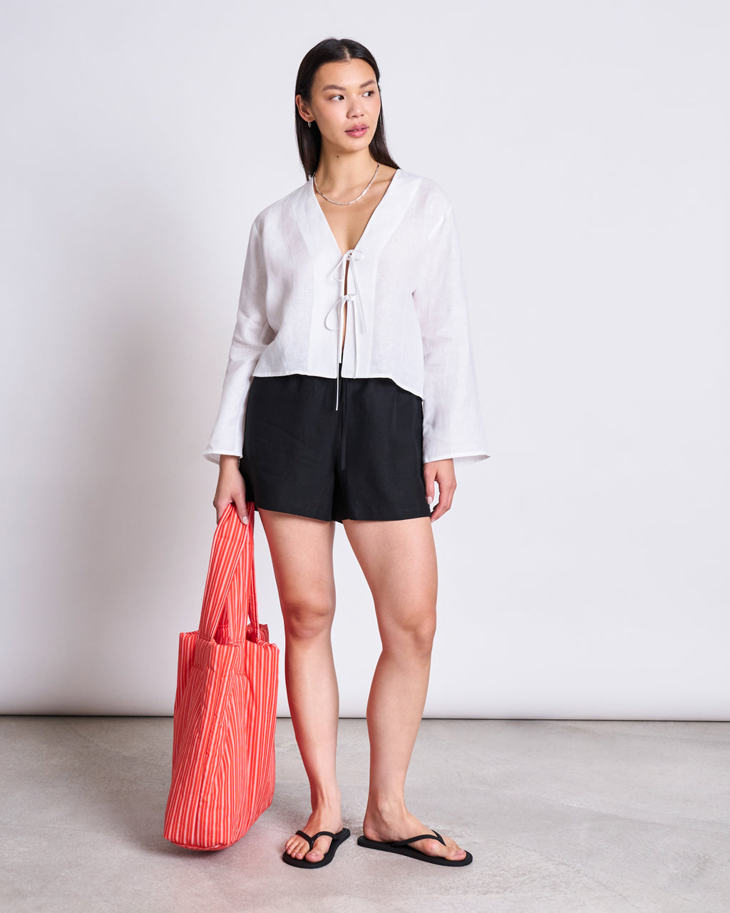 A person stands against a plain background wearing the jan-n-june LINEN TIE BLOUSE SKAGEN WHITE, paired with black shorts, black sandals, and holding a large red-striped tote bag.