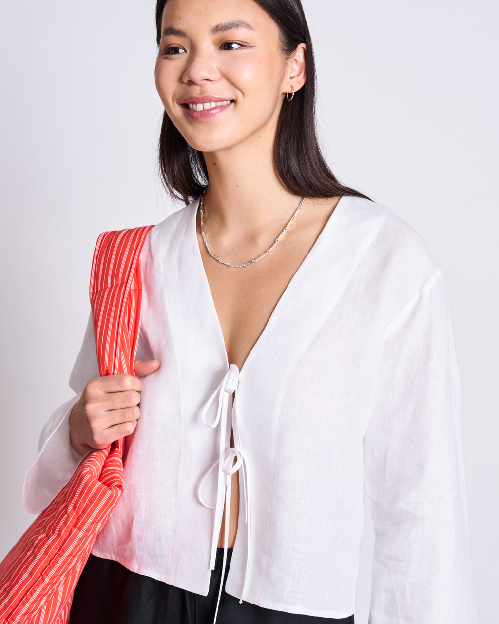 A woman smiles in the jan-n-june LINEN TIE BLOUSE SKAGEN WHITE with black pants, carrying a red-and-white striped tote and wearing a silver necklace, against a plain light background.