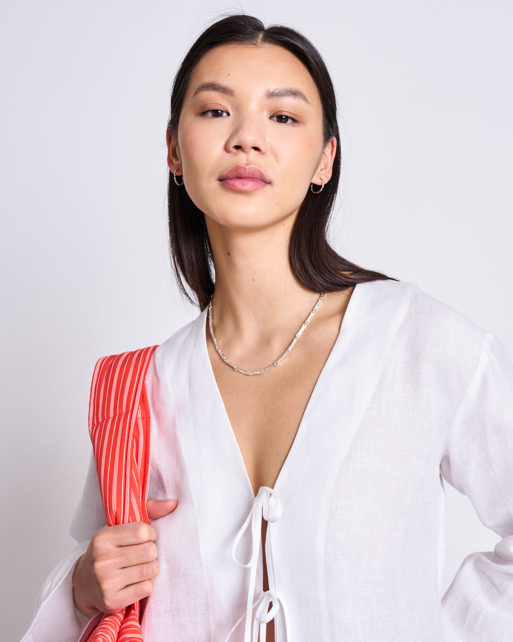 A woman with long dark hair wears the jan-n-june LINEN TIE BLOUSE SKAGEN WHITE, styled with a silver necklace and red-and-white striped bag, standing against a plain light background.