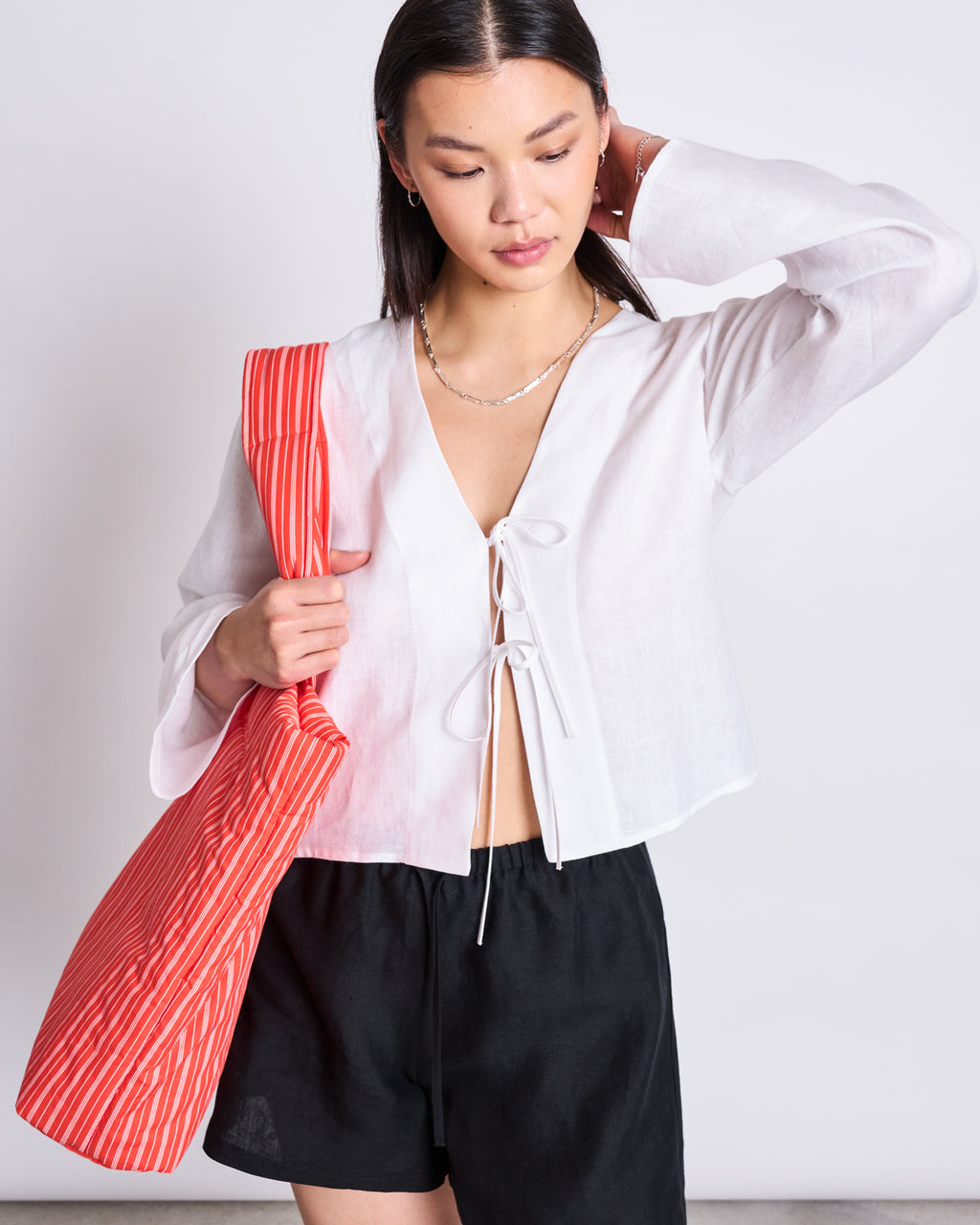 A woman wearing the jan-n-june LINEN TIE BLOUSE SKAGEN WHITE and black shorts stands holding a large red-and-white striped bag over her shoulder, looking down with a neutral expression.