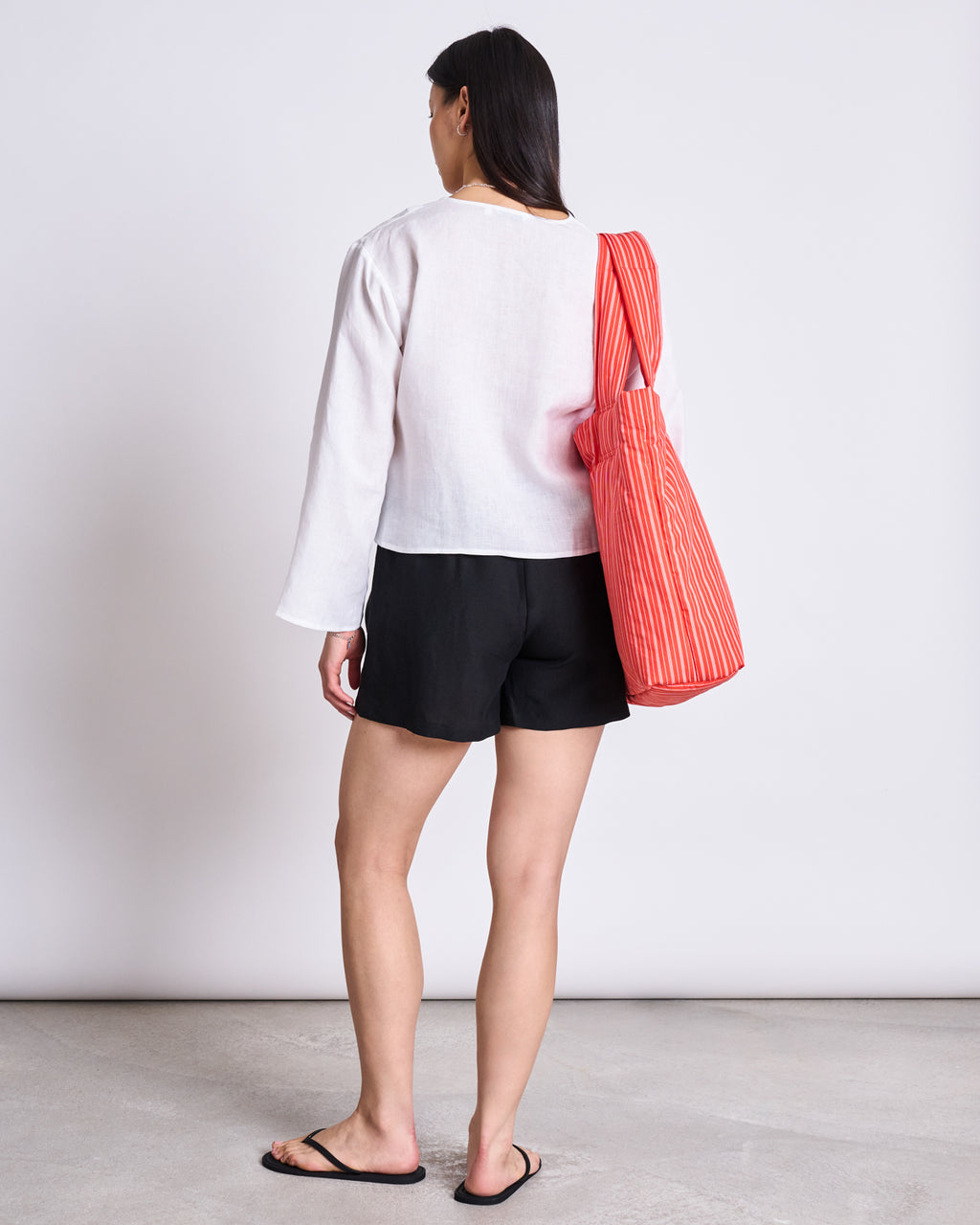 A person with straight dark hair, wearing the jan-n-june LINEN TIE BLOUSE SKAGEN WHITE, black shorts, and black flip-flops stands facing away with a red-and-white striped tote bag over their shoulder against a plain background.