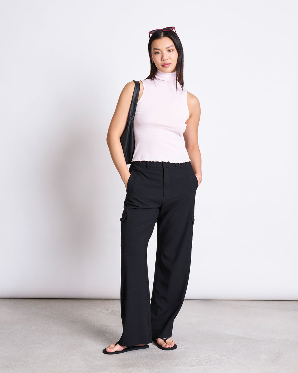 A woman stands against a white background, wearing a sleeveless light pink turtleneck, jan-n-junes CARGO PANTS FANO BLACK, black flip-flops, sunglasses on her head, and a black shoulder bag. Her hands are in her pockets.