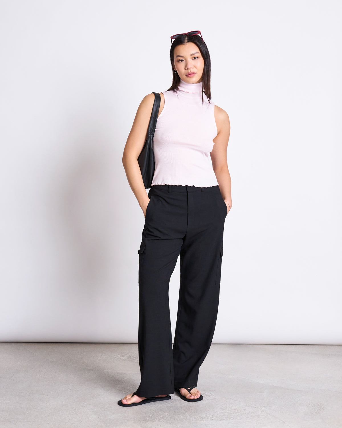 A woman stands against a white background, wearing a sleeveless light pink turtleneck, jan-n-junes CARGO PANTS FANO BLACK, black flip-flops, sunglasses on her head, and a black shoulder bag. Her hands are in her pockets.