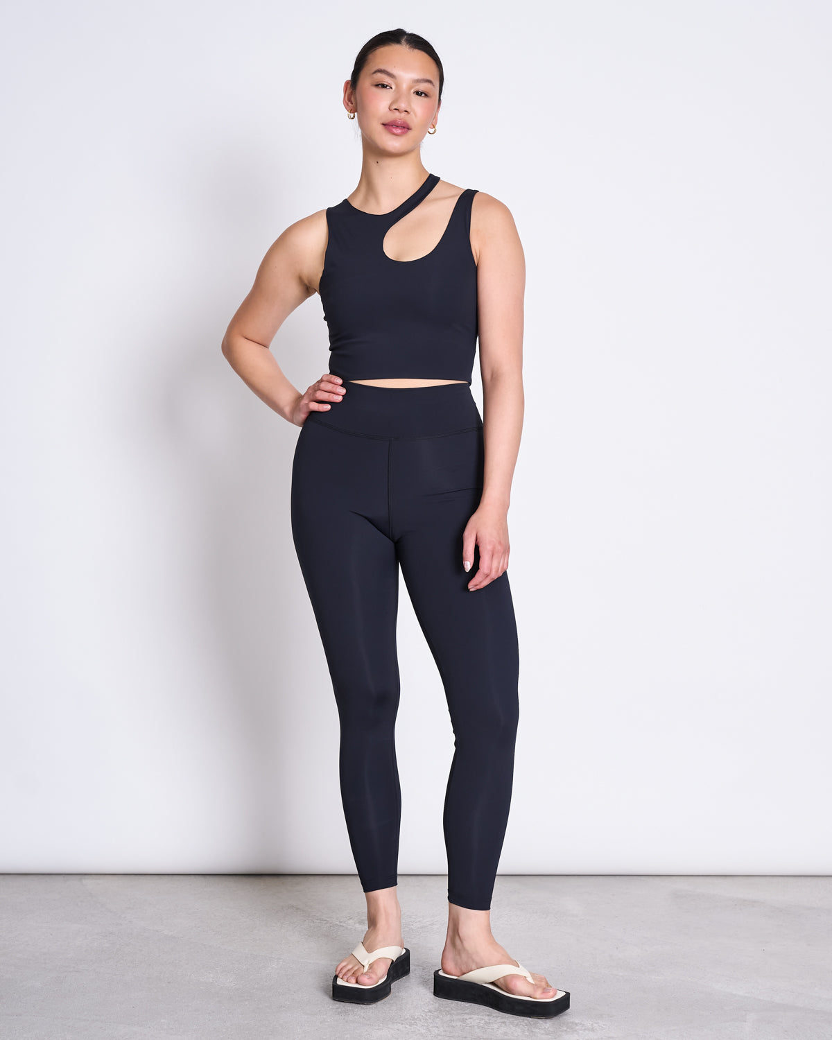 A woman with her hair tied back stands against a plain background wearing jan-n-june’s black crop top with a cutout and ACTIVE LEGGINGS HALLIN BLACK in recycled polyamide, paired with white flip-flop sandals. One hand rests on her hip.