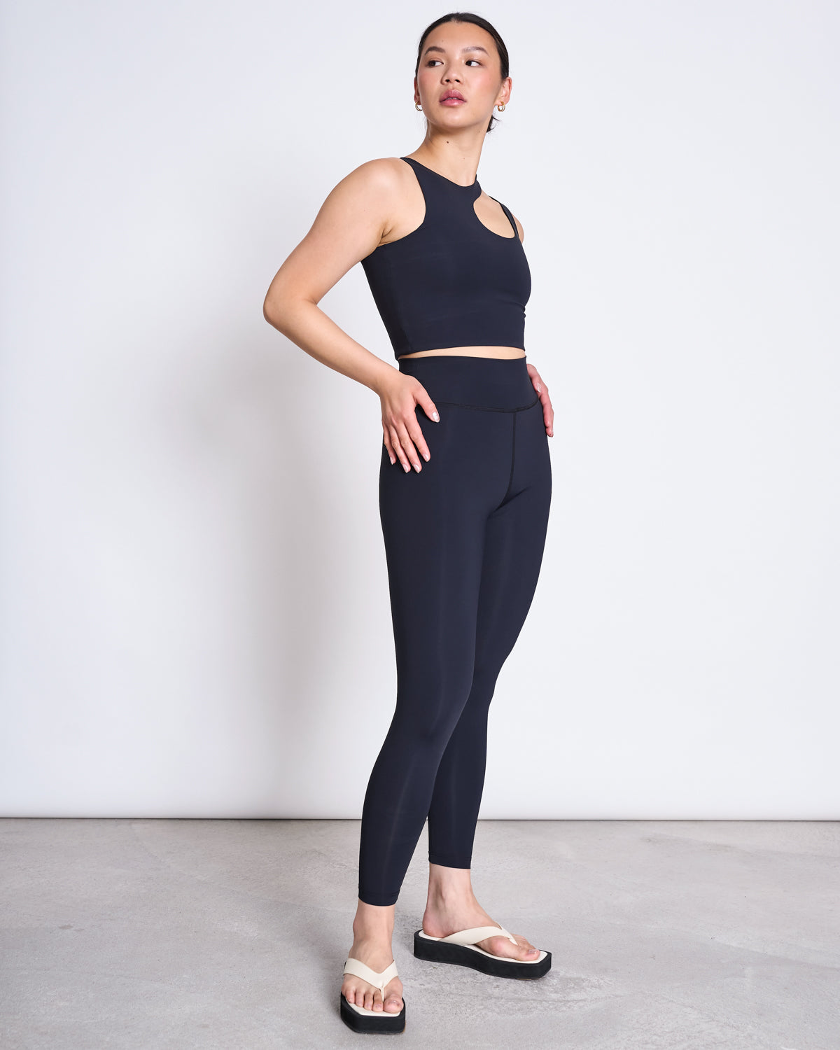 A person stands on a plain floor against a white wall, wearing a black sleeveless crop top and jan-n-june ACTIVE LEGGINGS HALLIN BLACK made from recycled polyamide, paired with beige sandals with black soles, looking to the side with one hand on their hip.
