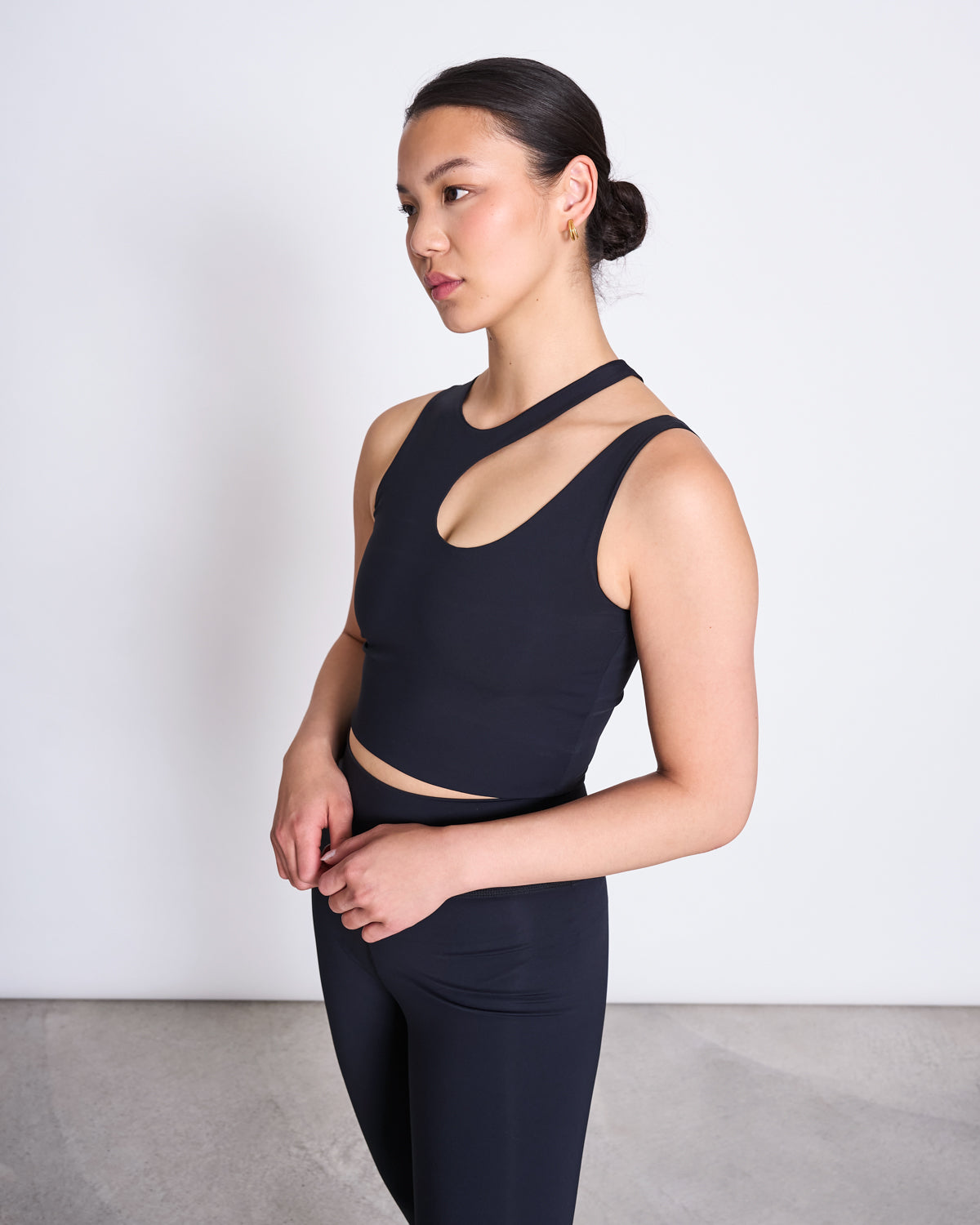 A woman with dark hair in a low bun wears a black sleeveless top with an asymmetric neckline and jan-n-june ACTIVE LEGGINGS HALLIN BLACK, made from recycled polyamide, standing against a plain white background.