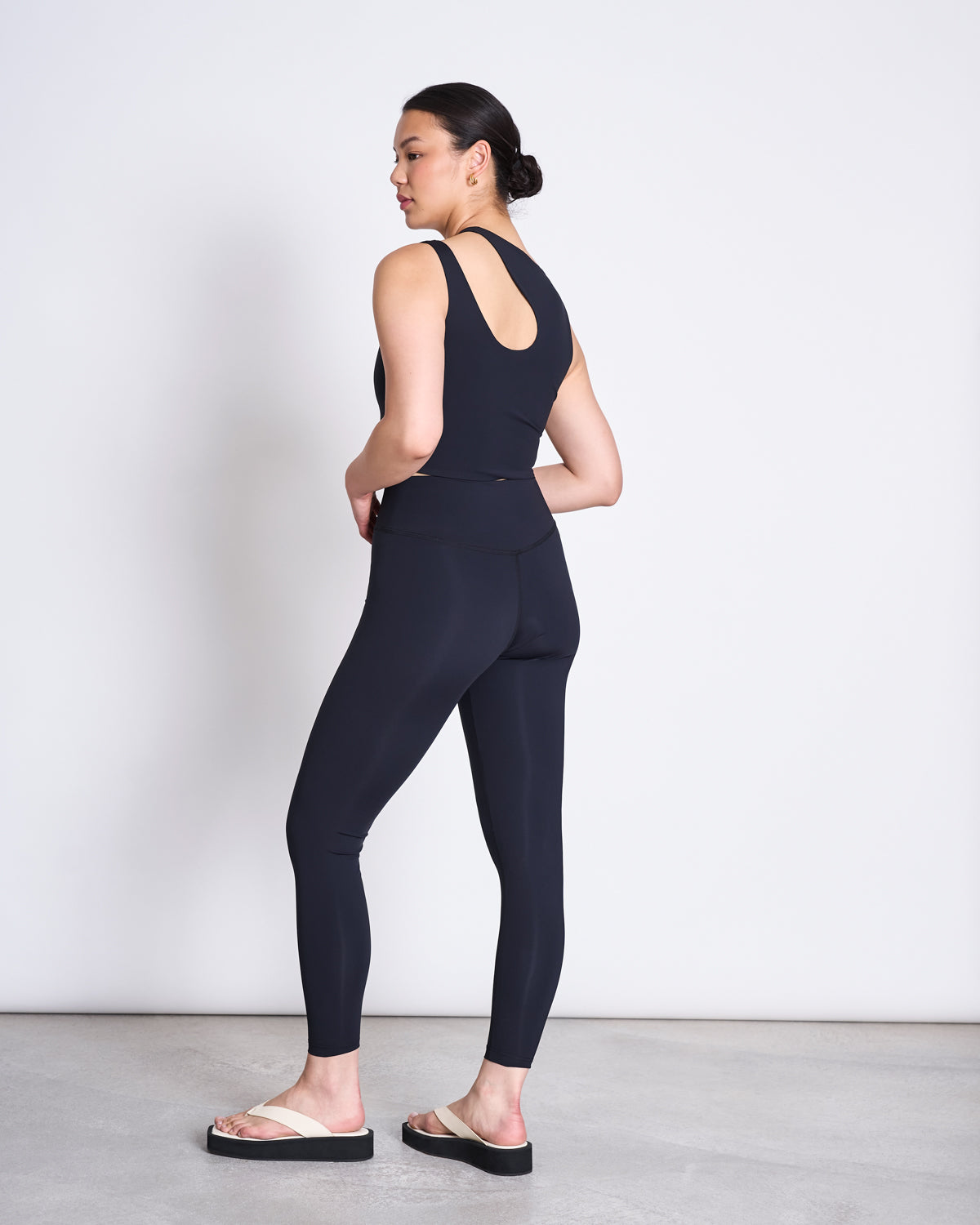 A woman in a minimalist room wears a black sleeveless top and jan-n-june ACTIVE LEGGINGS HALLIN BLACK, crafted from recycled polyamide with a neoprene-like finish, paired with white sandals. She looks over her shoulder, hair tied back.